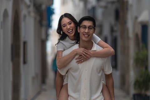 Happy couple enjoying a piggyback ride during a professional photoshoot in a scenic, narrow street in Sitges, Spain.